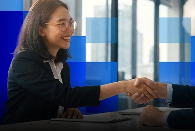 Two business professionals shake hands across a desk.