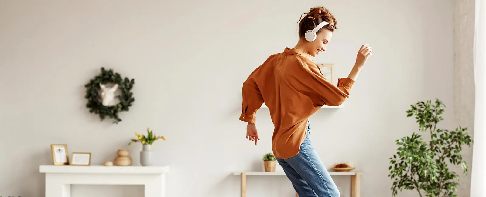 A person in a brown shirt dances joyfully in a cozy, bright room adorned with plants and decorative elements.