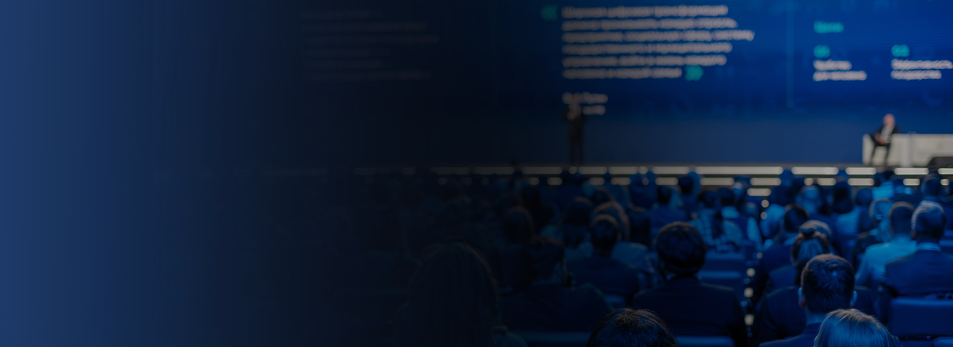 Delegates seated in a darkened conference hall, faces lit by a wide stage screen.