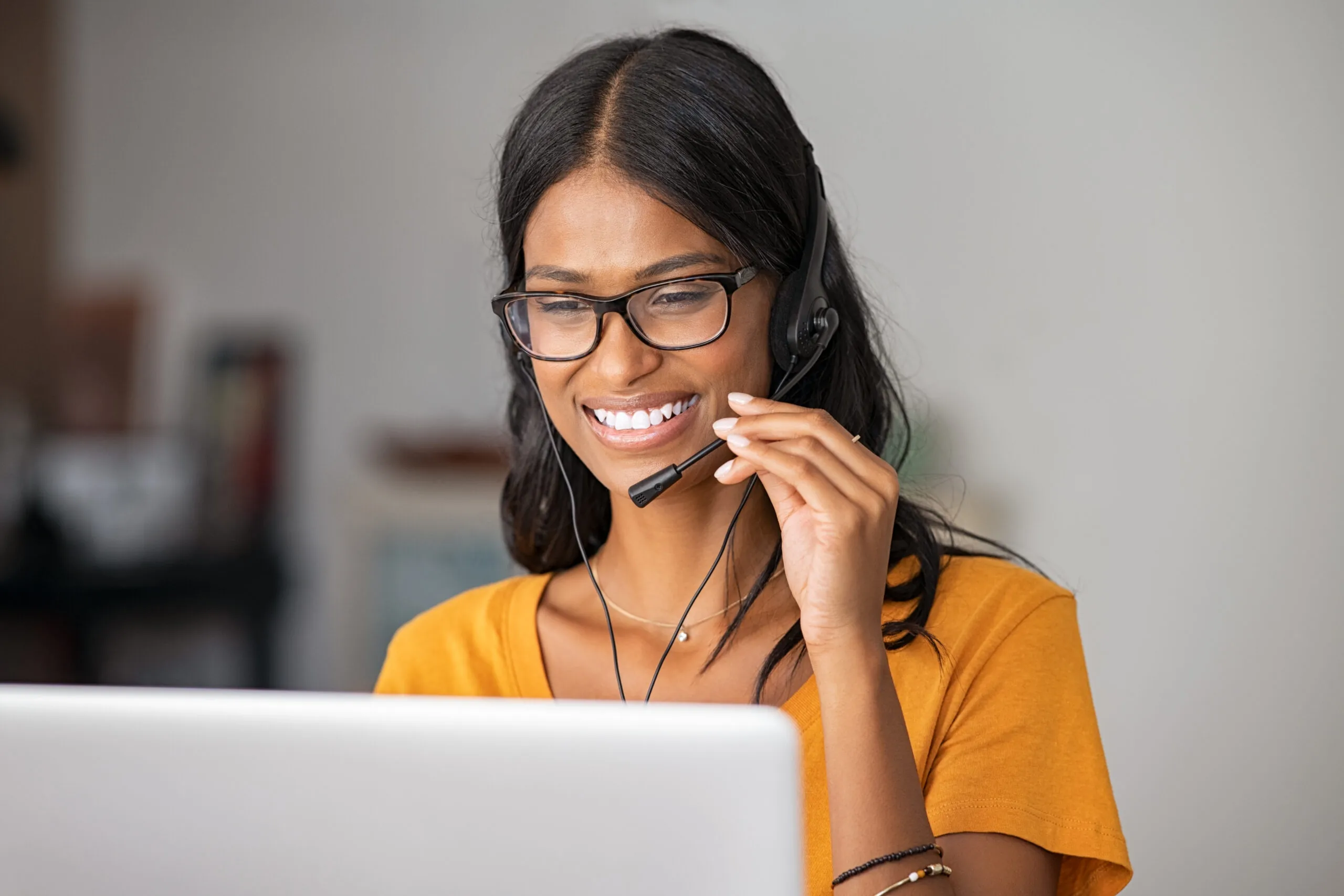 A woman in a yellow shirt wears a headset and speaks into a microphone while working at a computer.