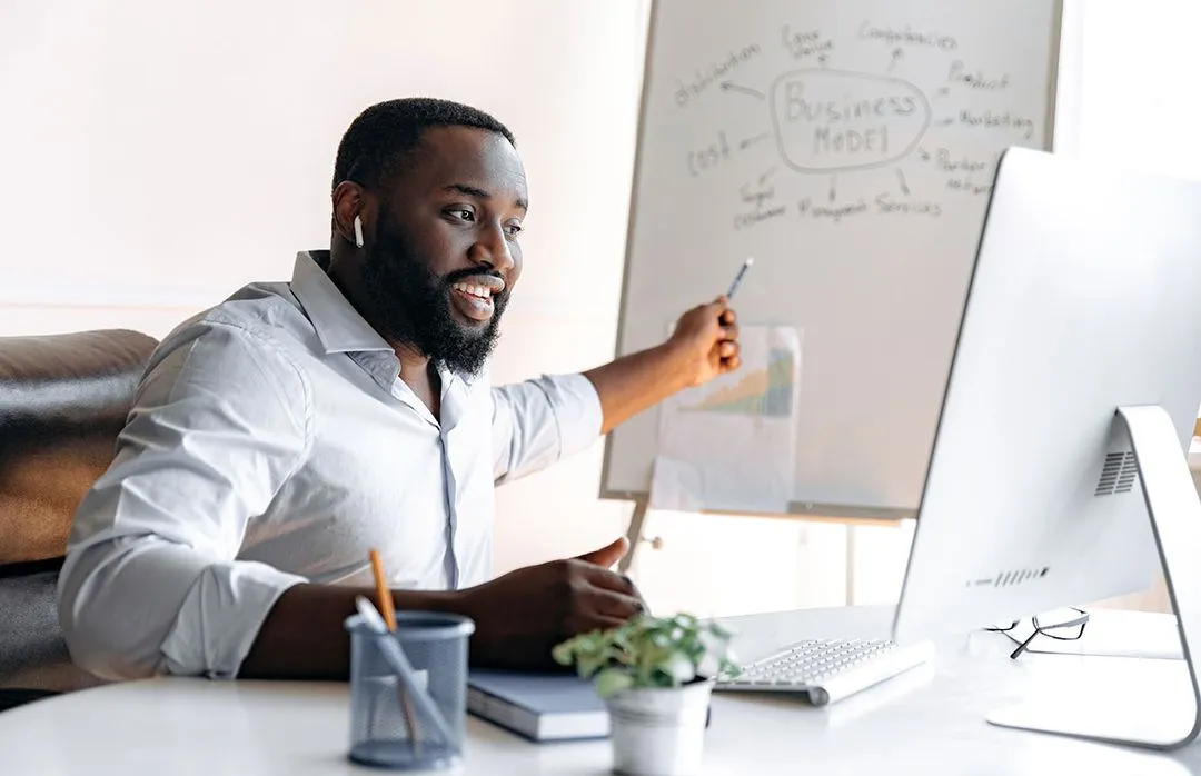 A business professional points at a whiteboard displaying a business model diagram, while seated at a modern desk with a computer.
