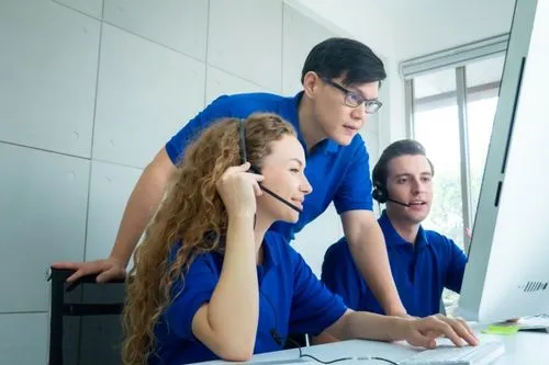 Three individuals in blue shirts collaborate around a computer, discussing work intently in a bright, modern office setting.