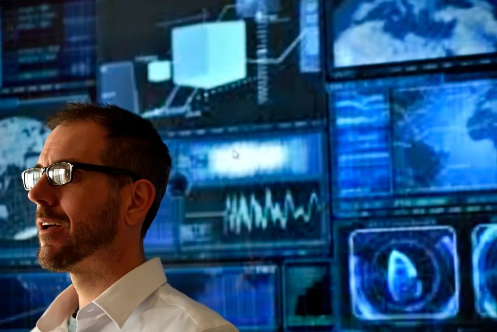 A person stands in front of a large, high-tech display featuring graphs, maps, and data visualizations in blue tones.