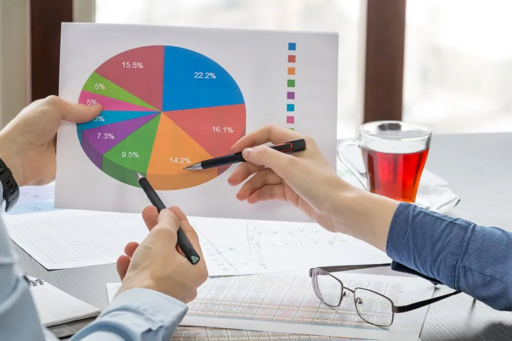 Two people analyze a color-coded pie chart on paper, discussing data with pens, while a cup of tea sits nearby.