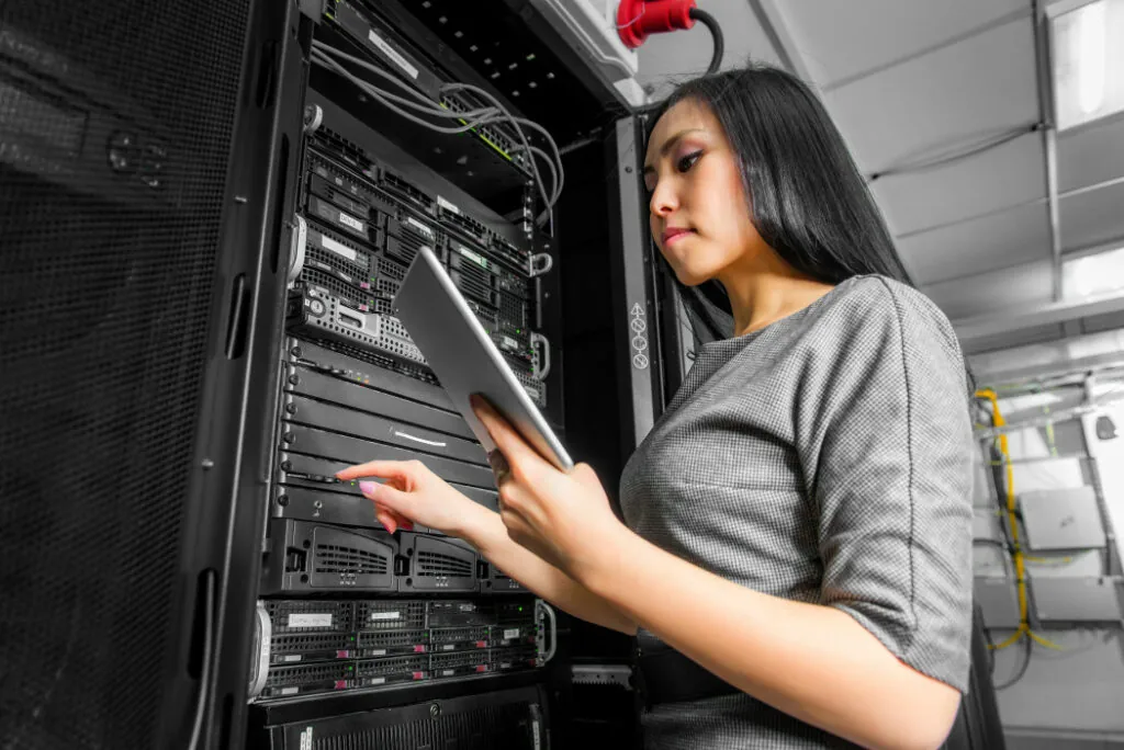 A person interacts with server equipment in a data center, holding a tablet and adjusting components in a digital environment.