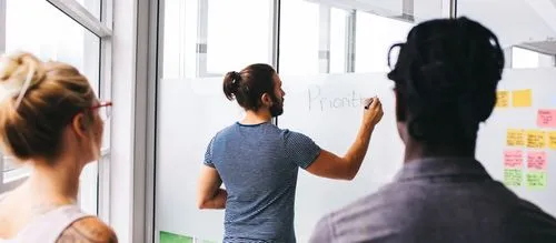 A group of three colleagues observes a man writing "Prioritize" on a glass board filled with colorful sticky notes.