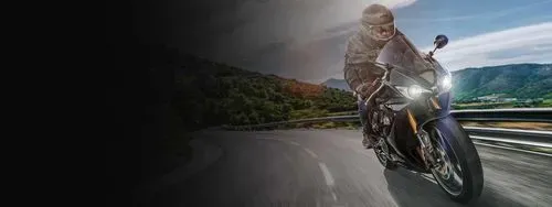 A motorcyclist in gear accelerates along a winding road, surrounded by lush green hills under a cloudy sky.