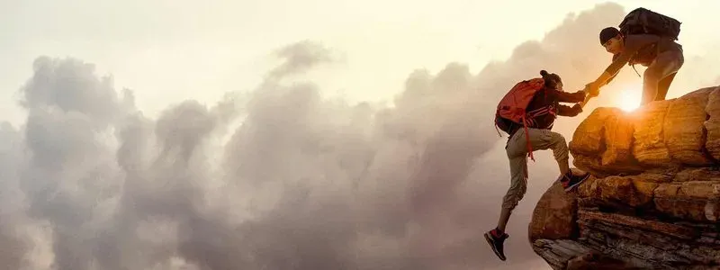 Two climbers assist each other on a rocky ledge at sunset, surrounded by clouds, showcasing teamwork and adventure.