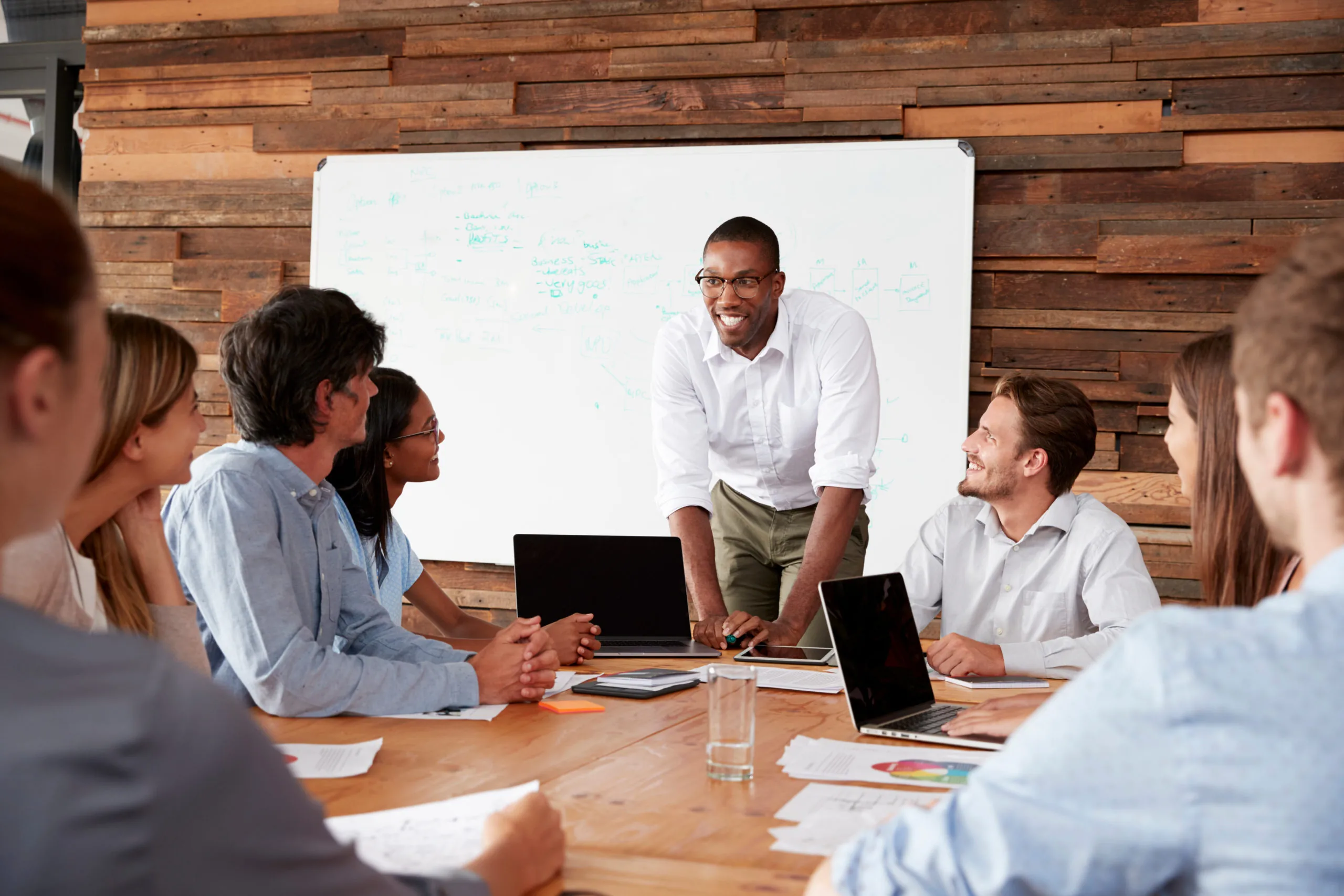 A diverse group of professionals engaged in a meeting, focusing on a presenter at a wooden table with laptops and a whiteboard.