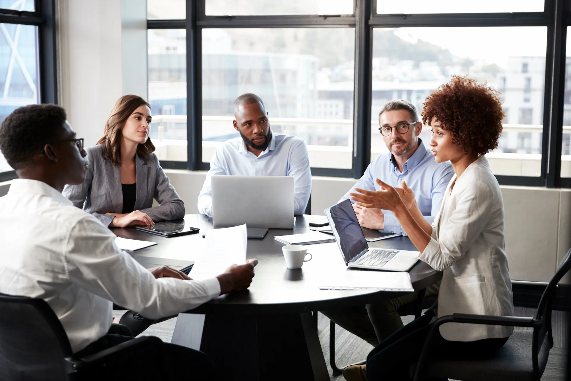 A group of professionals engaged in a collaborative meeting around a table, with laptops and documents, set in a bright office environment.