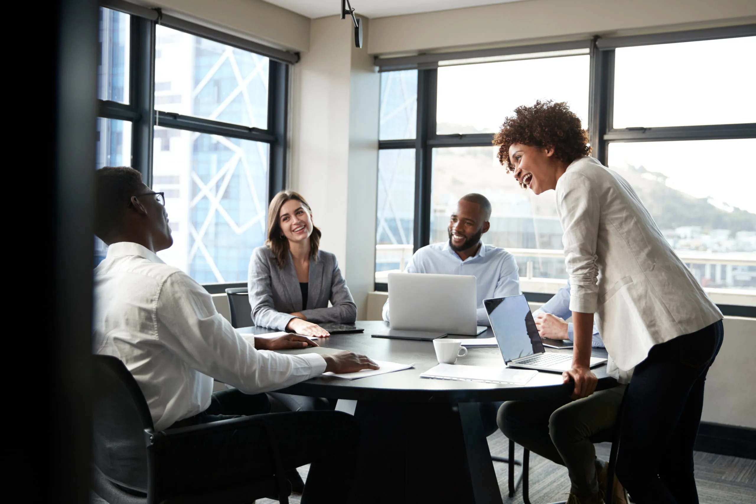 A diverse group of professionals engaged in a meeting around a conference table, with laptops and documents present. Natural light streams in through large windows.
