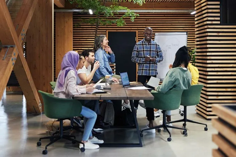 A diverse group of people collaborates around a table in a modern workspace, discussing ideas with laptops and notes.