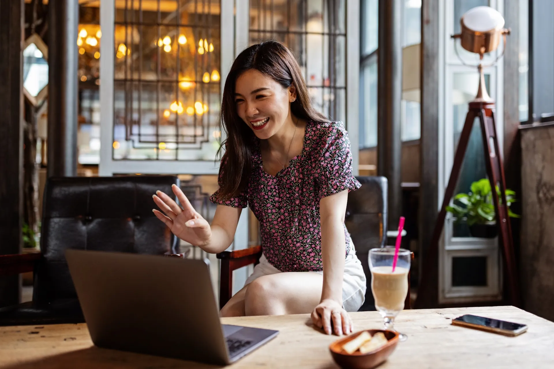 A woman waves to a laptop camera from a café table.