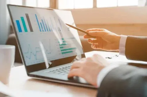 A business professional using a laptop to analyze charts and graphs, pointing with a pen in a bright, modern workspace.