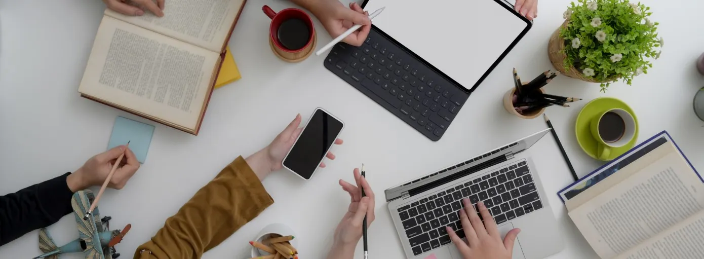 A top-down view of a workspace with hands using a laptop, tablet, smartphone, and notebooks, surrounded by coffee and plants.