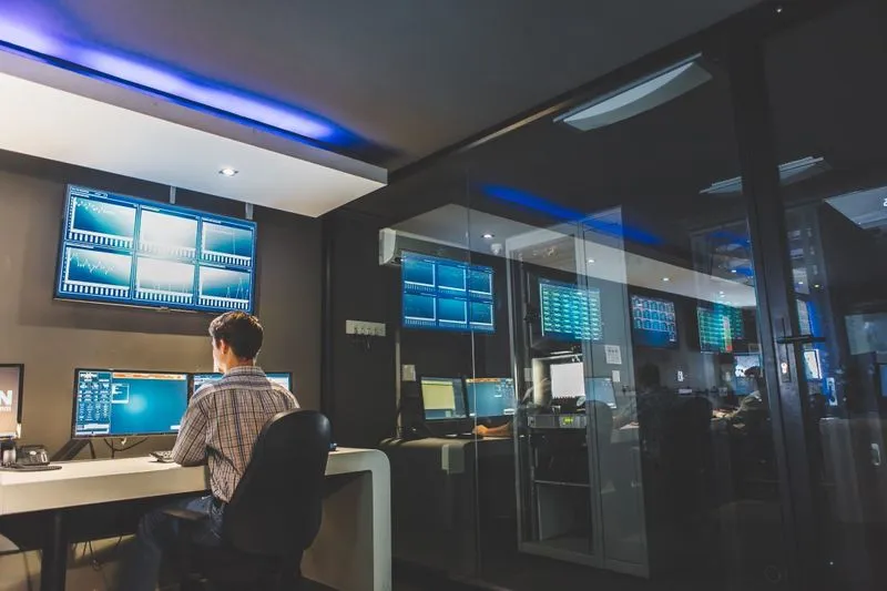 A man works at a computer in a dark office filled with multiple screens displaying data and graphs, showcasing a busy trading environment.