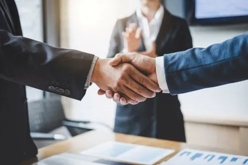Two business professionals shake hands in a modern office setting, while a third person stands in the background, applauding.
