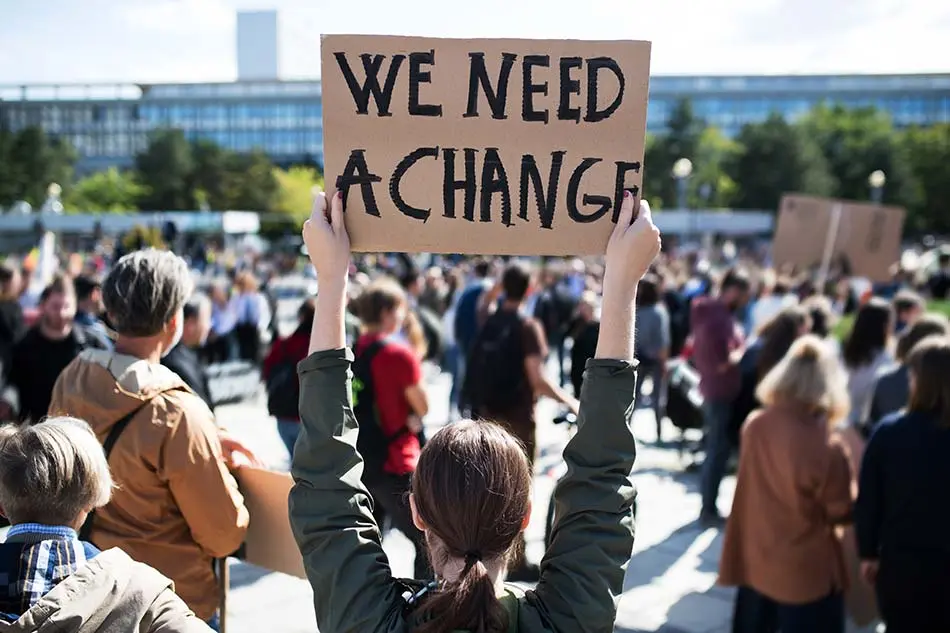A demonstrator at a crowded outdoor rally holds a hand-lettered cardboard sign reading “We need a change”.