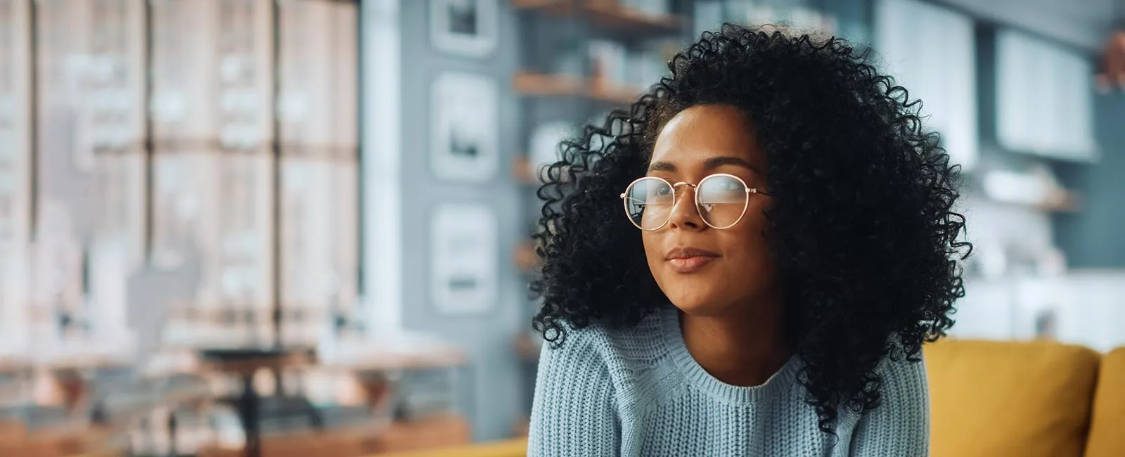 A person with curly hair wearing glasses sits on a couch in a stylish, cozy room with bookshelves and large windows in the background.