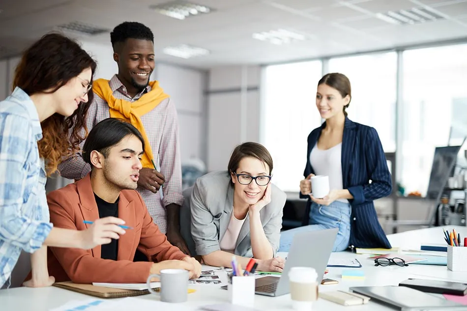 Five colleagues gathered around a desk in a bright open-plan office, looking together at an open laptop.