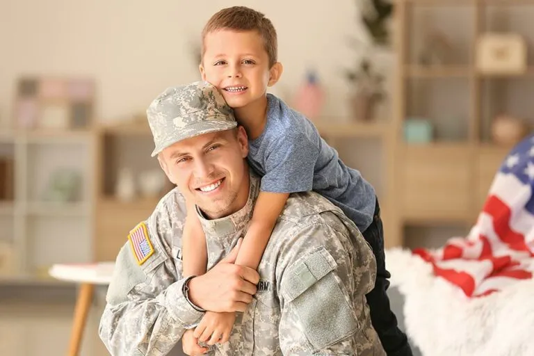 A military parent embraces a young child, both smiling, in a cozy living room with American decor in the background.