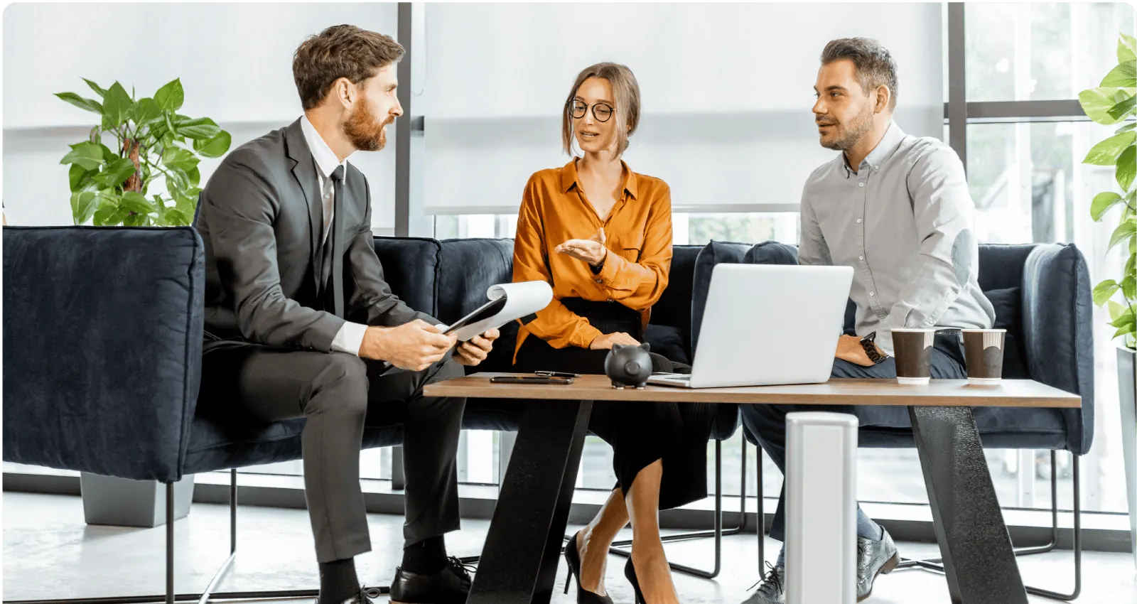 Three business professionals engaged in a discussion around a table with a laptop, coffee cups, and plants in a modern office setting.