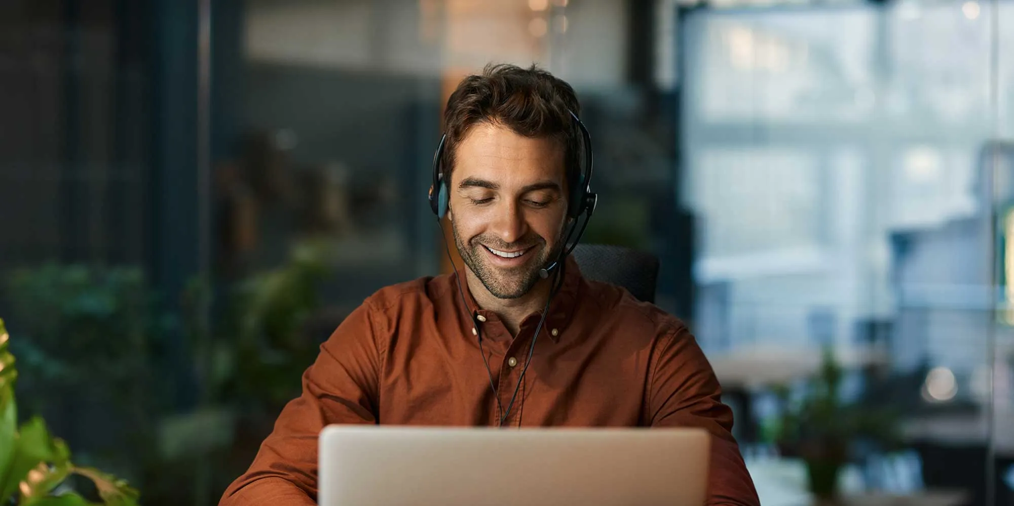 A man wearing headphones and an orange shirt is smiling while using a laptop.