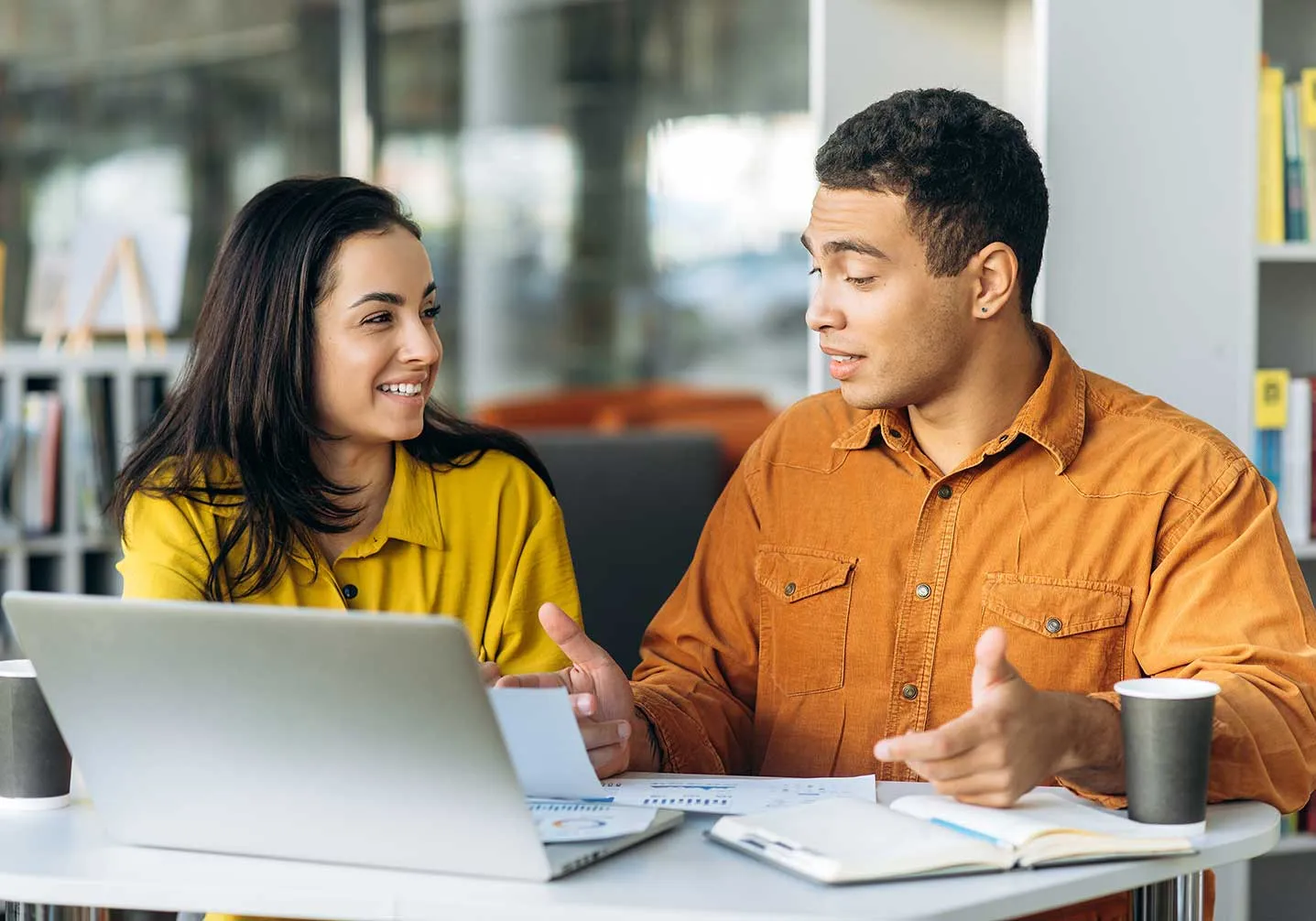 A woman in a yellow shirt and a man in an orange shirt engage in a discussion over a laptop and documents at a cozy table.