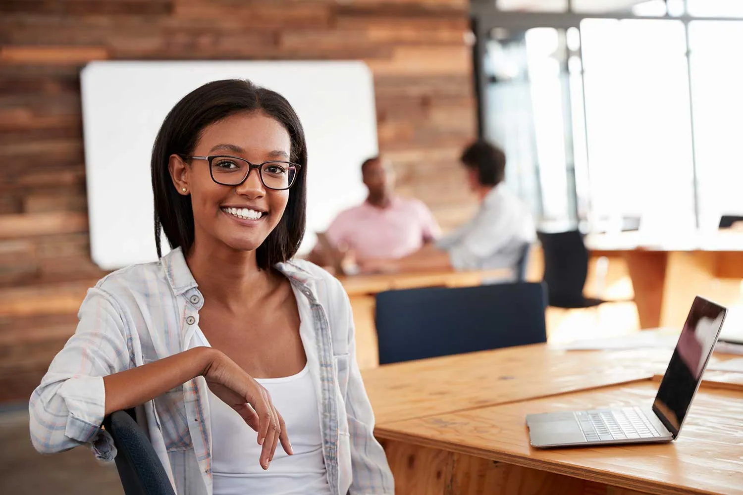 A young woman with glasses sits at a wooden table in a modern office, while colleagues converse in the background. A laptop is nearby.
