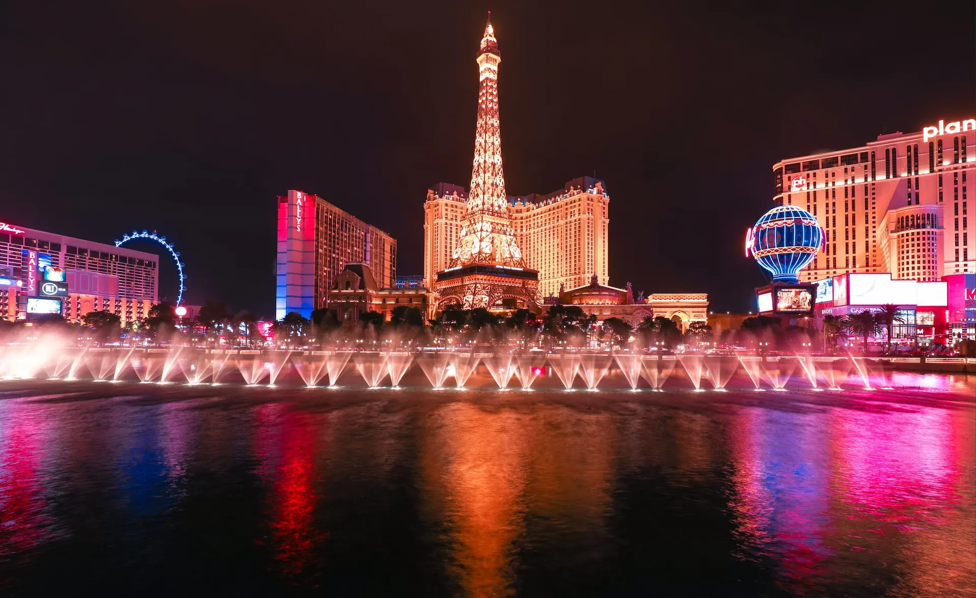 The Las Vegas Strip at night, featuring the Eiffel Tower replica, colorful lights, and a fountain display reflecting vibrant hues.