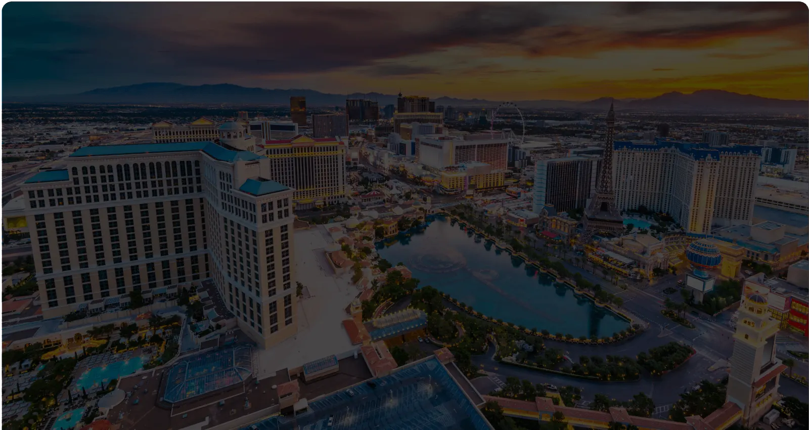 An aerial view of the Las Vegas Strip at dusk, with the Bellagio and its fountain lake in the foreground and the Eiffel Tower replica and High Roller wheel beyond.