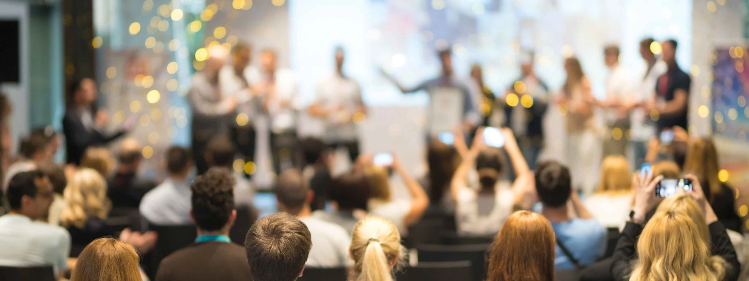 A crowd of attendees claps and takes photos as speakers are recognized on stage during a lively conference event.