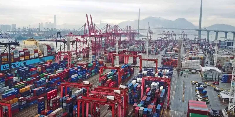 Aerial view of a busy cargo port filled with colorful shipping containers and cranes, with a city skyline and mountains in the background.