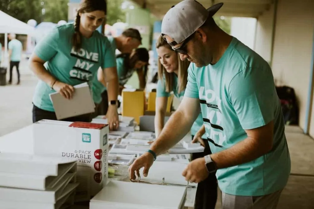 Volunteers in matching teal shirts organize supplies on a table during an outdoor community event, promoting a sense of collaboration and service.