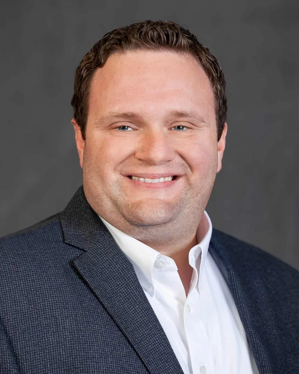 A man in a navy blazer and white shirt poses against a gray backdrop, with a professional demeanor.