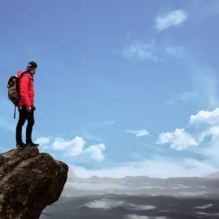 Hiker in a red jacket stands atop a rocky outcrop, gazing at a vast landscape under a bright blue sky with scattered clouds.