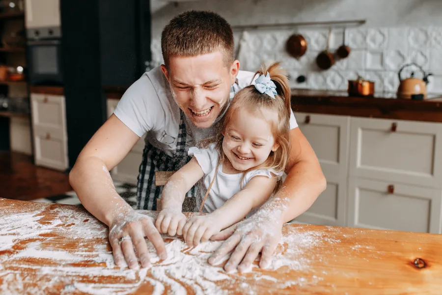 A parent and child share a joyful moment, playing with flour on a wooden kitchen table, creating a lighthearted baking scene.