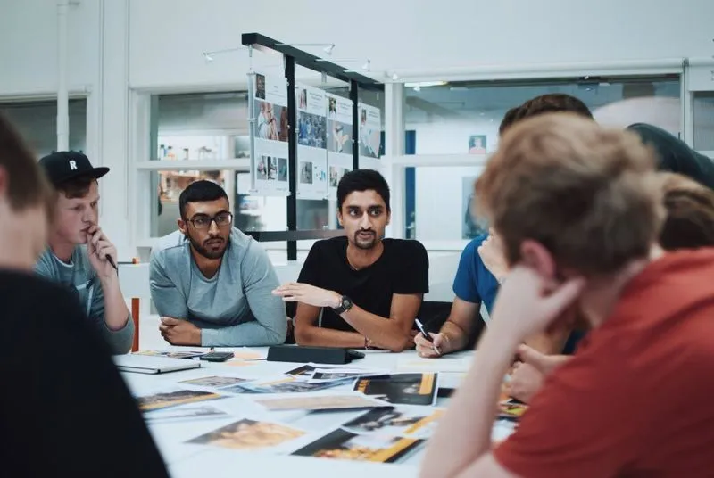 A group of diverse individuals engages in a collaborative discussion around a table covered with design materials and notes.