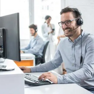 A person wearing headphones sits at a desk, typing on a computer, with another person in the background engaged in conversation.
