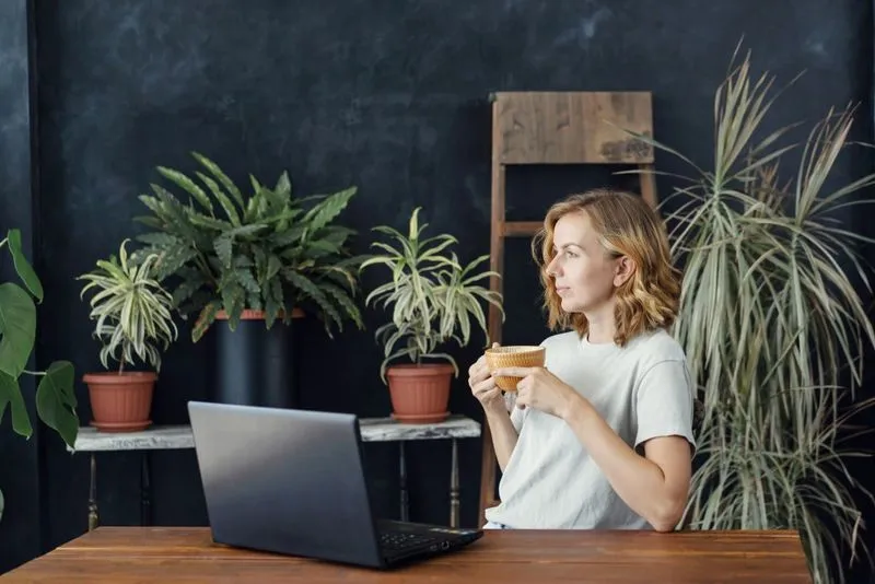 A person in a white shirt sits at a wooden table, holding a cup, with a laptop and potted plants in a cozy workspace.