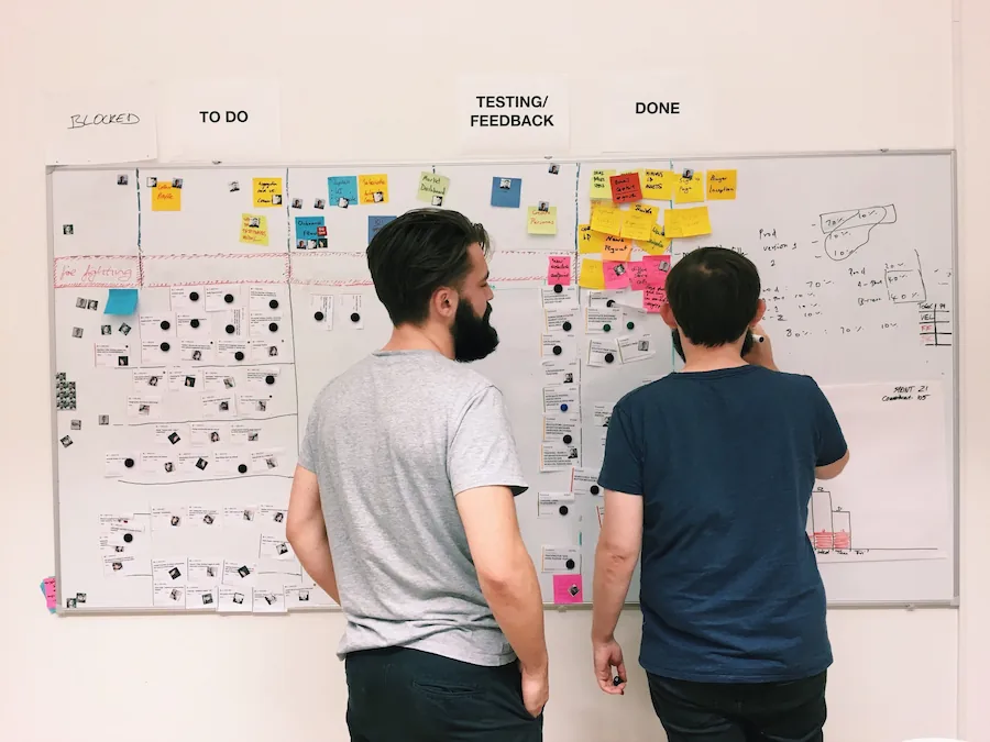 Two individuals reviewing a project board filled with sticky notes, charts, and progress indicators labeled "To Do," "Testing/Feedback," and "Done."
