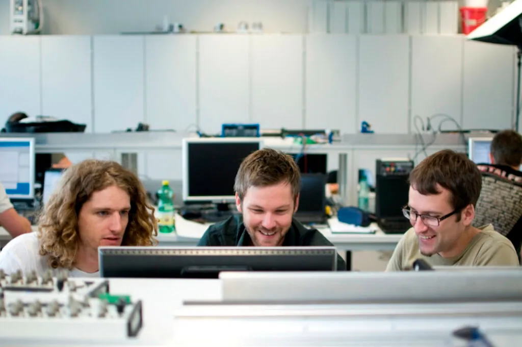 Three individuals focus on a computer screen in a tech workspace, surrounded by monitors and electronic equipment.