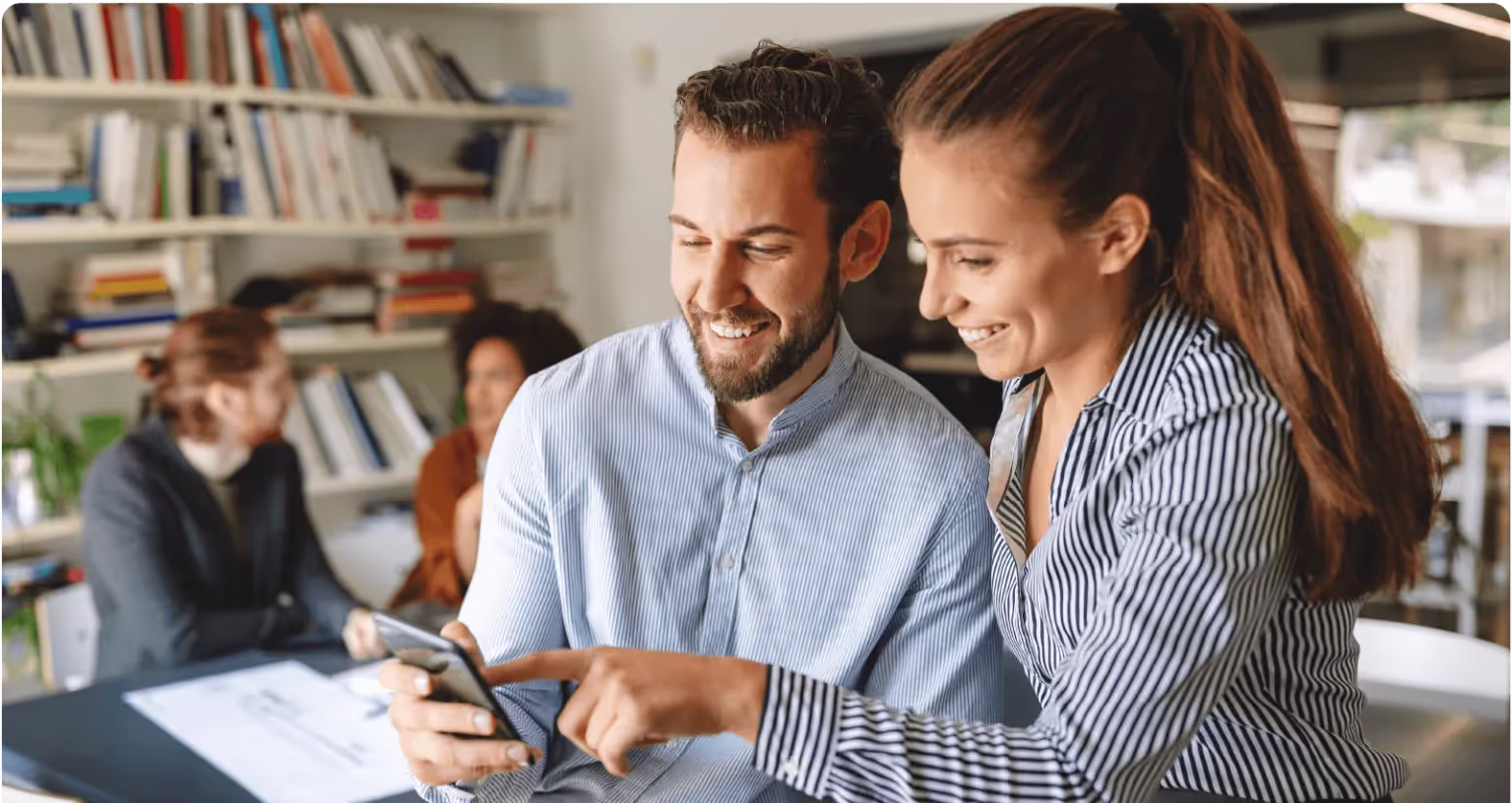 Two colleagues at a desk look at a phone together and smile, with two more people working behind them.