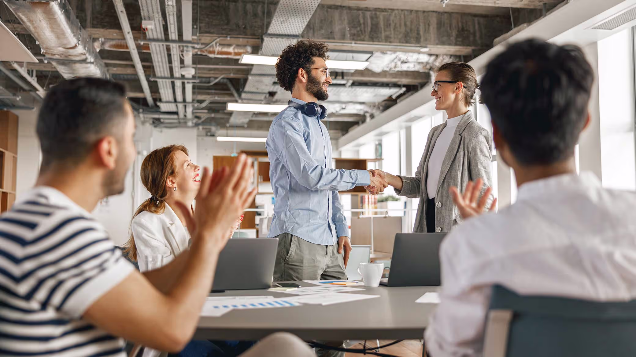 Two people shake hands across a meeting table in an open-plan office while three seated colleagues applaud.