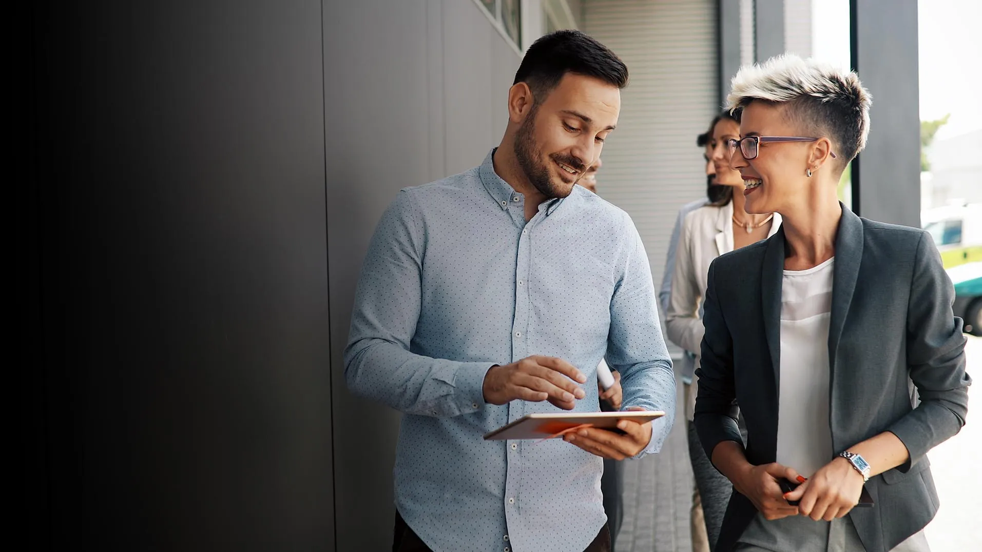 Two colleagues walk through a modern office corridor reviewing a tablet together, with two more of their team behind them.