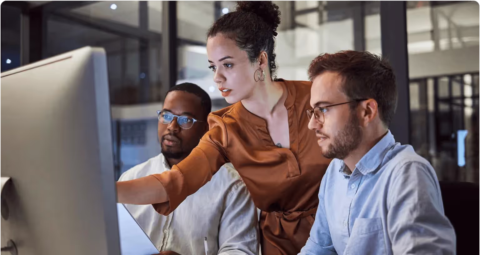 Three colleagues gathered at a desktop monitor, one of them pointing something out on the screen.