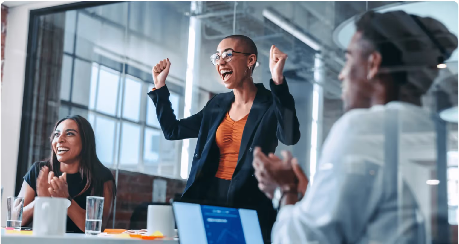 A woman stands at a meeting table with both fists raised in celebration while two colleagues applaud.