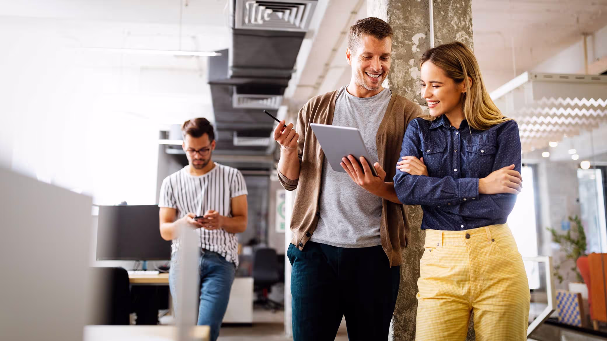 Two colleagues stand together in an open-plan office looking at a tablet, with a third working on a phone behind them.