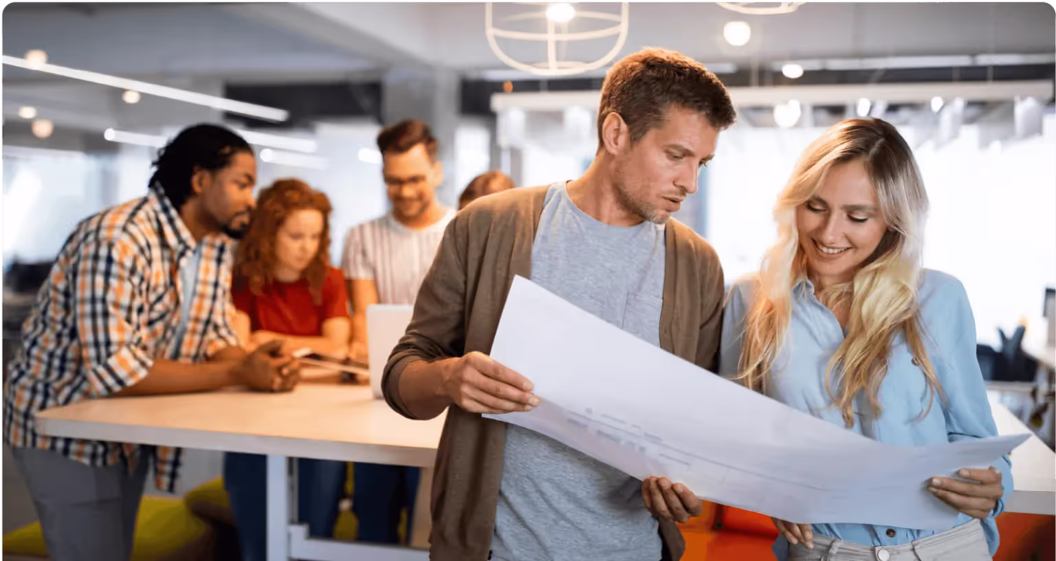 Two colleagues study a large unrolled printed plan in an open-plan office while others work at a table behind them.
