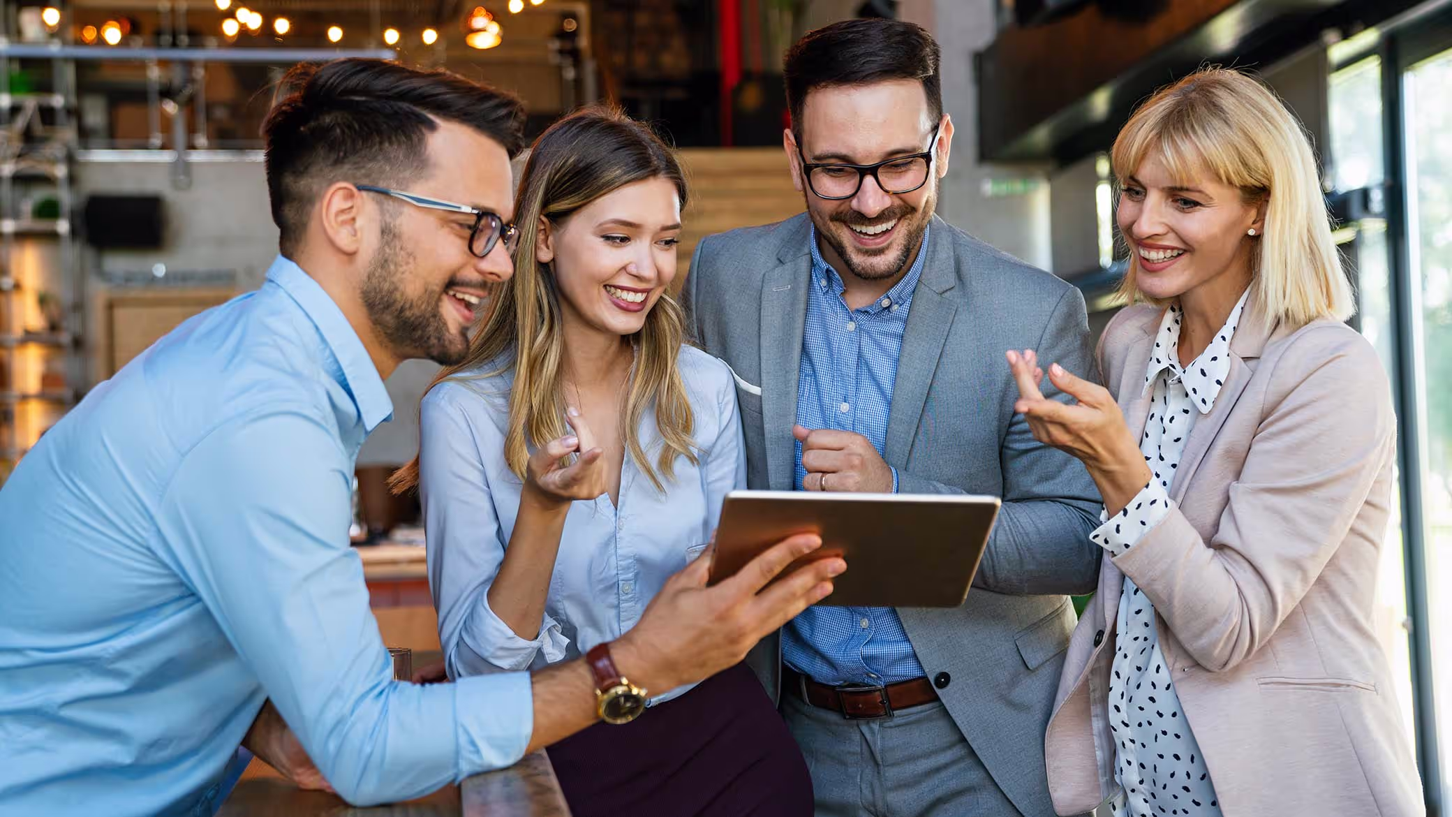 Four colleagues stand together looking at a tablet one of them is holding, smiling and talking.